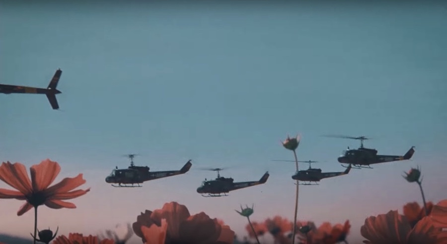 A pink flower in the foreground with multiple helicopters flying in the sky in the background.