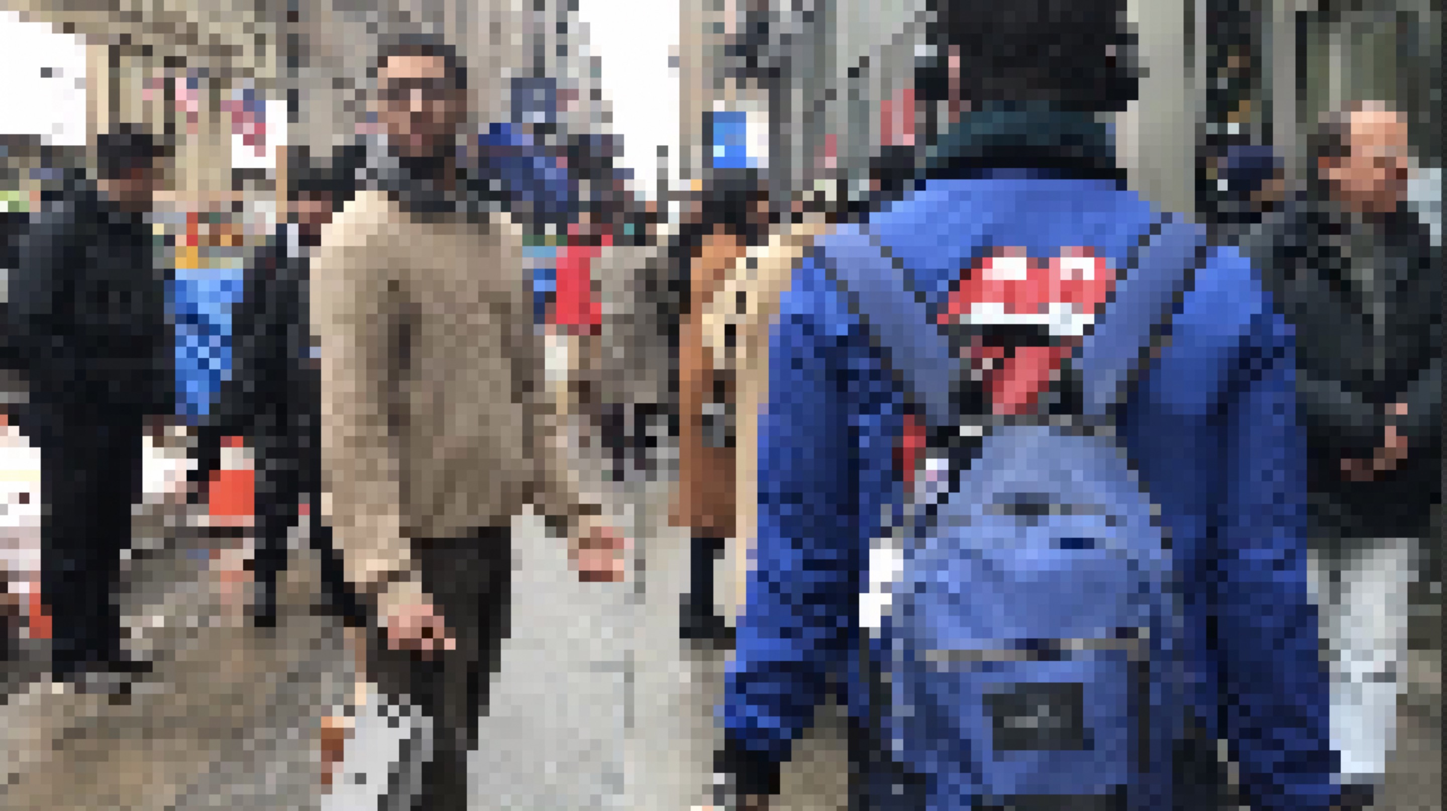 A pixelated photo outside on the streets of Midtown Manhattan. A man in a beige sweater in the center frame looks toward the camera. Someone with a blue jacket with lips on it and a blue backpack walks away from the camera.